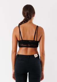 Bralette in pizzo nero con spalline regolabili, caratterizzato da un pannello trasparente e un motivo floreale, abbinato a pantaloni neri a vita alta con tasche posteriori.
