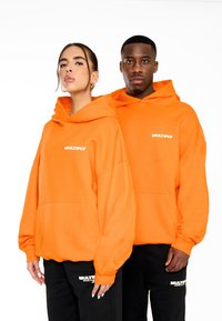 Sudaderas naranjas de gran tamaño con bolsillos frontales y puños de canalé, que presentan un logo blanco de "MULTIPLICAR" en el pecho. Pantalones negros con acentos de logo.