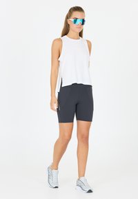 Top bianco senza maniche con orlo curvo, abbinato a pantaloni corti neri a vita alta con tasche laterali. Indossato con occhiali da sole riflettenti blu e sneakers grigie.
