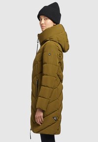 Giacca a vento in verde oliva, con collo alto, design a zig-zag trapuntato, cappuccio e tasche laterali con zip. Abbinata a un berretto in maglia nero.