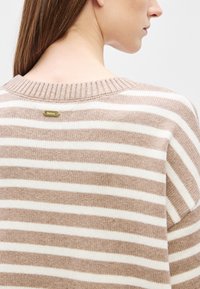 Vöödiline kootud kampsun helepruunide ja valgete triipudega, soonikuga kaelusega ja väike kuldne logomärk taga. Pehme tekstuur.