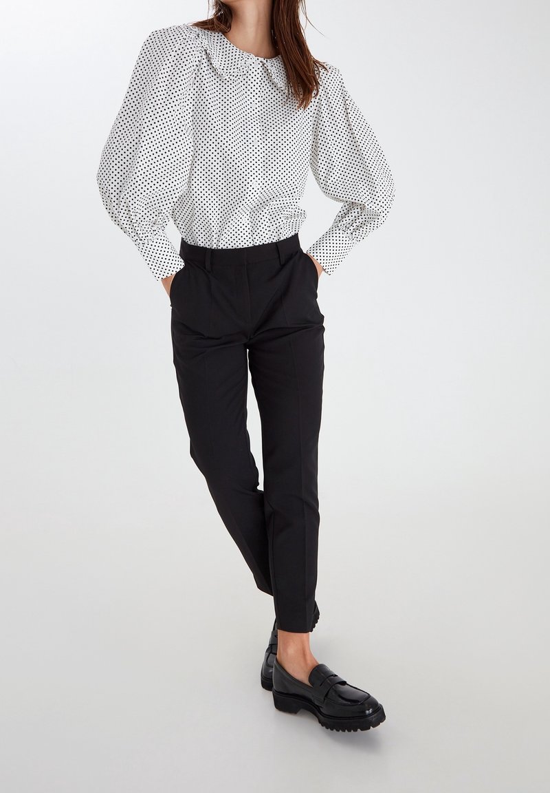 Donna che indossa una camicia bianca con pois neri e maniche a sbuffo, pantaloni ankle neri su misura e mocassini neri, in piedi con le mani nelle tasche.