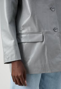 Sacou din piele gri cu o textură netedă, cu două butoane și un detaliu de buzunar singular. Asortat cu blugi denim de culoare albastră deschisă la baza.