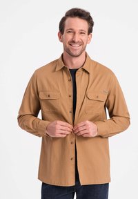 Beige button-up shirt met twee voorkruiken, gemaakt van een stevig fabric. Heeft een kraag en een rechte zoom. Donkere onderhemd.