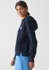 Felpa blu navy con un fit rilassato, tasca a marsupio e logo frontale. Materiale morbido con polsini e orlo a coste, dotata di un dettaglio con apertura laterale.