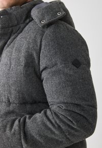 Pilkas pūkinis džemperis su siuvinėtu dizainu, aukštu apykakle ir snapelių užsegimu. Audinys turi tekstūrinį, melžtinį paviršių.