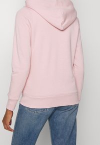 Persona che indossa un felpa con cappuccio rosa chiaro e jeans in denim blu, vista da dietro su uno sfondo semplice.