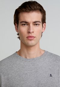 Grijze katoenen t-shirt met een ronde halslijn en een klein geborduurd doodskop en kruisbotten logo op de linkerkant van de borst. Gladde textuur.
