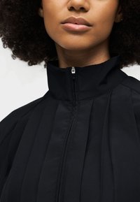 Nahaufnahme einer Person, die eine schwarze Jacke mit hohem Kragen und Reißverschluss vorne trägt, zeigt das untere Gesicht und strukturiertes lockiges Haar.