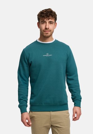 Felpa verde acqua con scollo rotondo, caratterizzata da un logo bianco sul petto, polsini a coste e tessuto strutturato. Abbinata a pantaloni beige.