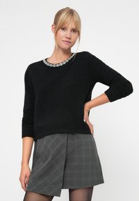 Black knit sweater with a round neckline featuring a decorative silver embellishment. Model paired it with a gray plaid skirt.