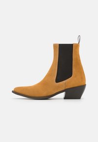 Suede ankelsko i tan med en svart elastisk sidopanel, spetsig tå och en avsmalnande svart klack. Har synliga sömdetaljer.