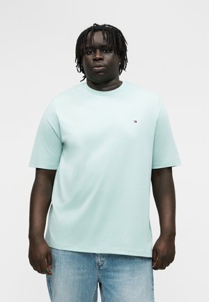 Άνδρας με σκούρο δέρμα και κοτσίδες dreadlocks, φοράει ανοιχτό μπλε κοντομάνικο πουκάμισο και ανοιχτόχρωμο τζιν παντελόνι, στέκεται μπροστά σε απλό φόντο.