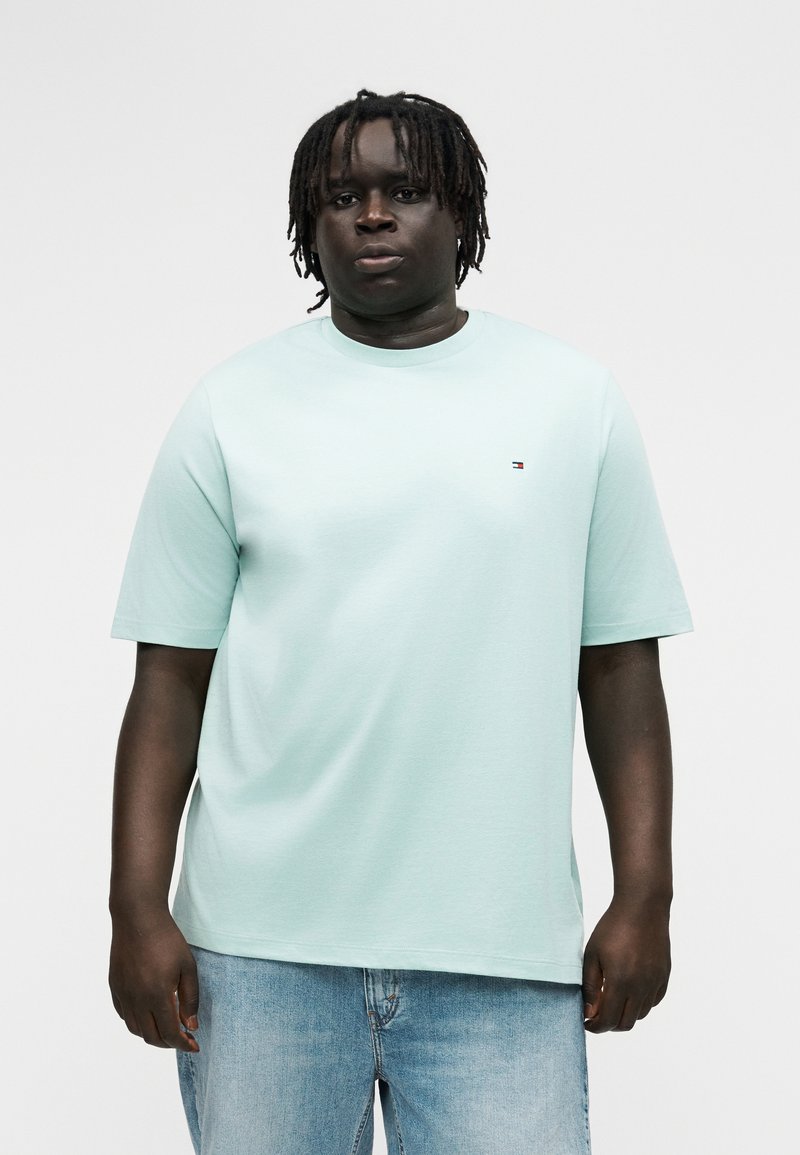 Άνδρας με σκούρο δέρμα και κοτσίδες dreadlocks, φοράει ανοιχτό μπλε κοντομάνικο πουκάμισο και ανοιχτόχρωμο τζιν παντελόνι, στέκεται μπροστά σε απλό φόντο.