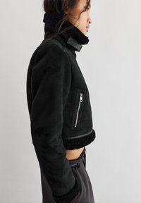 Giacca nera corta in morbido suede con colletto in shearling, tasca con zip e polsini testurizzati, caratterizzata da elementi di design minimalisti.