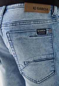 Jeans in denim azzurro chiaro con finitura testurizzata, dotati di un'etichetta in cuoio marrone sulla cintura e di un piccolo marchio nero sulla tasca posteriore.