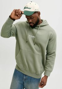 Olivgrön hoodie med framficka och dragsnören, med texten "JJ Studios" i vitt. Matchas med en keps i kräm och grönt.