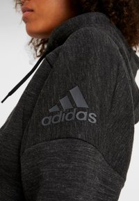 Felpa nera con texture e logo Adidas prominente in finitura opaca sulla spalla, con cappuccio regolabile e dettagli a contrasto.