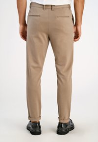 Calças chino bege com textura suave, apresentando dois bolsos traseiros e bainhas enroladas. Combinadas com sapatos de couro pretos.