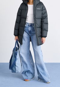 Černá péřová bunda s kapucí, žebrovanými manžetami a logem; bílý žebrovaný top; široké modré džíny; modrá kabelka z různých látek.