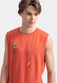 T-shirt sans manches orange en coton lisse. Présente des figures abstraites d'enfants et des flèches brodées de différentes couleurs.