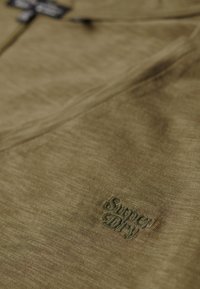 Oliven grøn bomulds sweatshirt med en struktureret finish, der har et broderet "Superdry"-logo i matchende grøn på brystet.