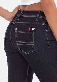 Dunkelblaue Denim-Jeans mit kontrastierendem gelben Nähten, zwei Gesäßtaschen und einem kleinen roten Logowimpel neben der rechten Tasche. Enganliegendes Design.