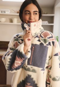 Pile con motivi geometrici in crema, verde, rosa e blu. Presenta un collo alto e una tasca sul petto con zip in tessuto a costine.