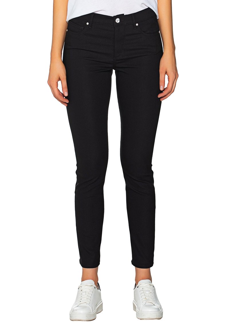 Femme portant un pantalon noir slim et des baskets blanches, debout, les bras détendus le long du corps, sur un fond uni.