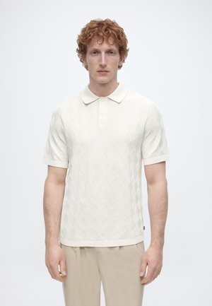 Man met krullend rood haar, gekleed in een wit textuur-korte mouwen poloshirt en beige broek, staand tegen een effen lichte achtergrond.