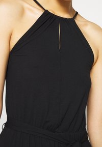 Gros plan d'une personne portant une robe noire sans manches à encolure américaine avec une petite découpe en forme de trou de serrure à l'avant et une ceinture nouée à la taille.