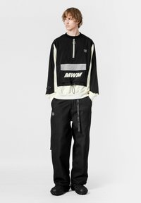 Chaqueta de viento negra con cuello con cremallera, detalles en blanco y un bolsillo delantero con el logo "MWM"; combinada con pantalones negros texturizados y calzado.