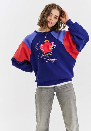 Kolorowy sweatshirt z niebieskim korpusem, czerwonymi i fioletowymi rękawami oraz graficznym wzorem przedstawiającym owoce i grzyby z napisem "Uważaj na słodkie rzeczy".