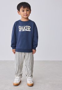 Marineblauwe trui met "BAKER"-logo, gecombineerd met crème gestreepte broek. Schoeisel heeft een wit bovenwerk met klittenbandsluitingen en een tankleurige zool.