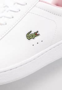 Sneaker bianca con logo di coccodrillo ricamato verde, dettagli tonali sottili e accenti rosa attorno al colletto.