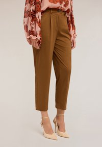 Un pantalon brun sur mesure présentant une texture lisse, une coupe fuselée et des détails plissés à l'avant, associé à des chaussures à talons beiges.