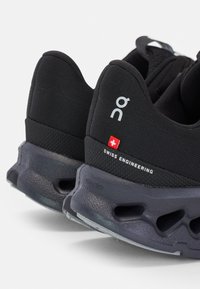Schwarze Sportchuhe verfügen über ein strukturiertes Obermaterial, eine Helion-Schaumsohle und eine Schweizer Ingenieur-Branding mit einem roten Schweizer Fähnchenakzent.