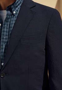 Navyblauer strukturierter Blazer mit taillierter Passform, einreihigem Knopfverschluss und einer Tasche. Getragen über einem blauen Karoshirt.