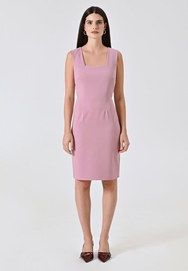 Rosa engangierte Kleid mit quadratischem Ausschnitt, ärmellosem Design und glatter Textur. Getragen mit spitzen braunen Absätzen, vor einem hellen Hintergrund.