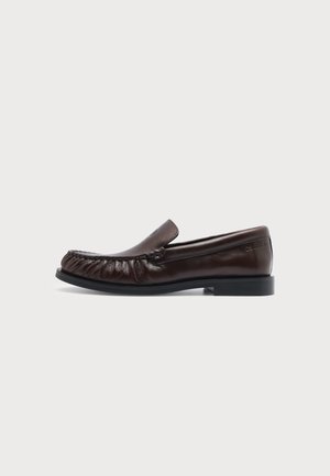 Mocassino in pelle marrone scuro con cucitura frontale raccolta e suola nera bassa, mostrato di profilo su uno sfondo chiaro uniforme.
