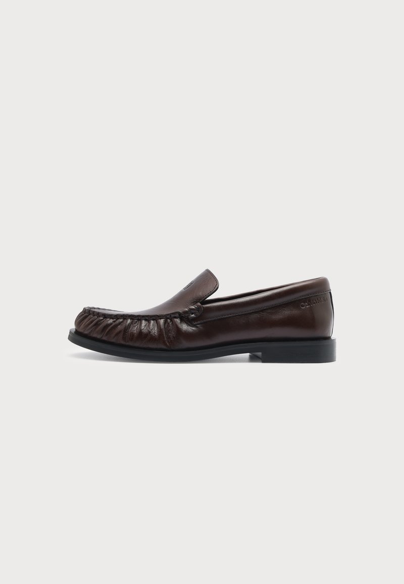 Mocassin en cuir marron foncé avec couture avant froncée et semelle basse noire, montré de profil sur un fond clair uni.