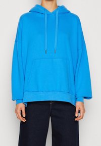 Felpa blu oversize in morbido tessuto, con tasca frontale, cappuccio regolabile e maniche larghe. Abbinata a jeans scuri.