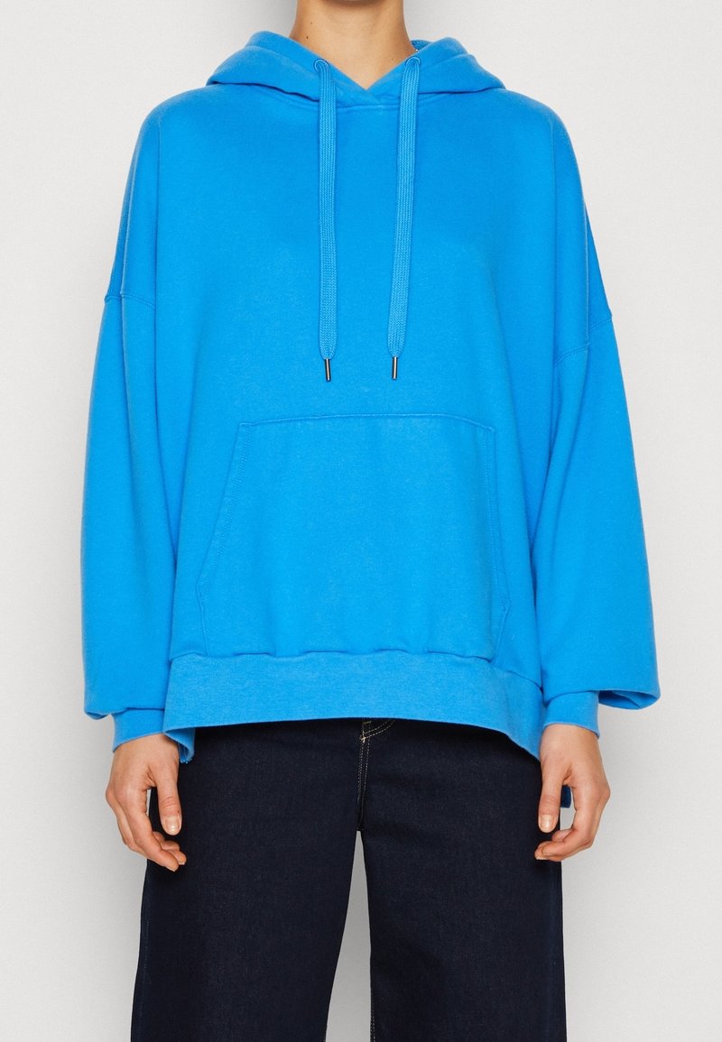 Felpa blu oversize in morbido tessuto, con tasca frontale, cappuccio regolabile e maniche larghe. Abbinata a jeans scuri.