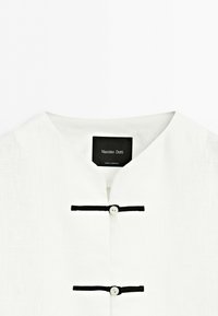 Bílá košile s mandarinkovým límečkem, dvěma černými horizontálními sponami se dvěma bílými knoflíky a viditelným štítkem Massimo Dutti.