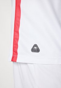 Camiseta atlética blanca con franjas laterales rojas, que presenta un logo triangular negro con el texto "KOMBAT SYSTEM" cerca del dobladillo. Textura suave.