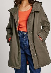 Oliwkowa parka z kapturem, z zamkiem z przodu, z dwoma bocznymi kieszeniami i pomarańczową podszewką. Połączona z jasnopomarańczową bluzką i niebieskimi dżinsami.