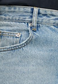 Τζιν παντελόνι ανοιχτού μπλε χρώματος σε κοντινό πλάνο που δείχνει την μπροστινή τσέπη με μεταλλικά καρφιά, θηλιά για ζώνη και κίτρινες ραφές.