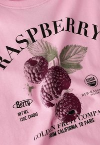 Chemise rose clair avec un graphique de framboises, texte noir "Framboise" et d'autres détails, avec des feuilles vertes et une étiquette biologique.