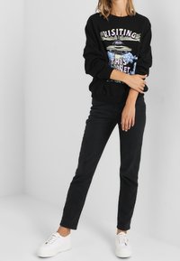 Zwart grafisch sweatshirt met kleurrijke tekst en ontwerp, gecombineerd met zwarte jeans en witte sneakers. Casual outfit op een neutrale achtergrond.