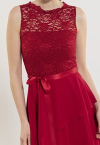Robe en dentelle rouge avec un haut sans manches et un décolleté festonné, ainsi qu'une jupe fluide ornée d'une ceinture en satin brillant nouée à la taille.